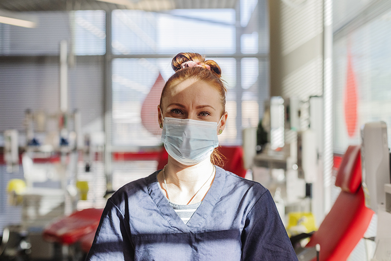 Veripalvelun hoitaja katsoo suoraan kameraan kasvoillaan maski.