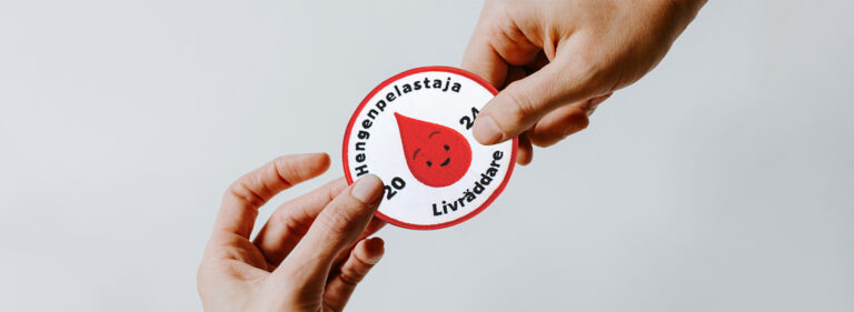 Kaksi kättä, jotka pitävät kiinni Hengenpelastaja-haalarimerkistä.