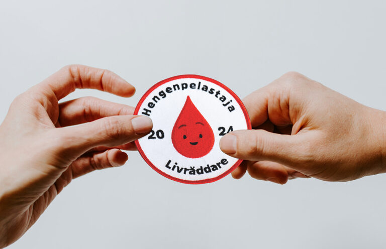 Kaksi kättä, jotka pitävät kiinni Hengenpelastaja-haalarimerkistä.