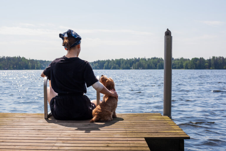 Ihminen istuu koiran kanssa laiturilla aurinkoisessa kesäsäässä.