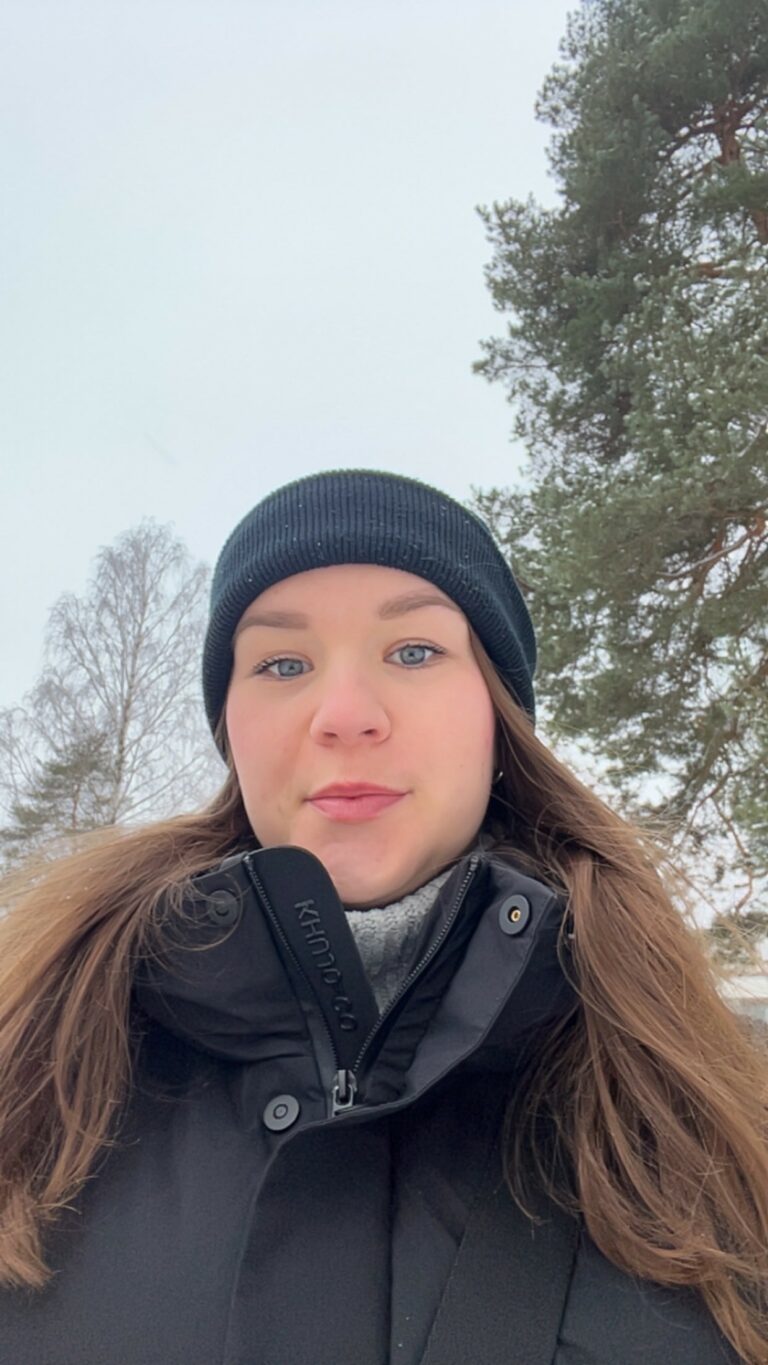 Nainen katsoo suoraan kameraan, taustalla näkyy puita.