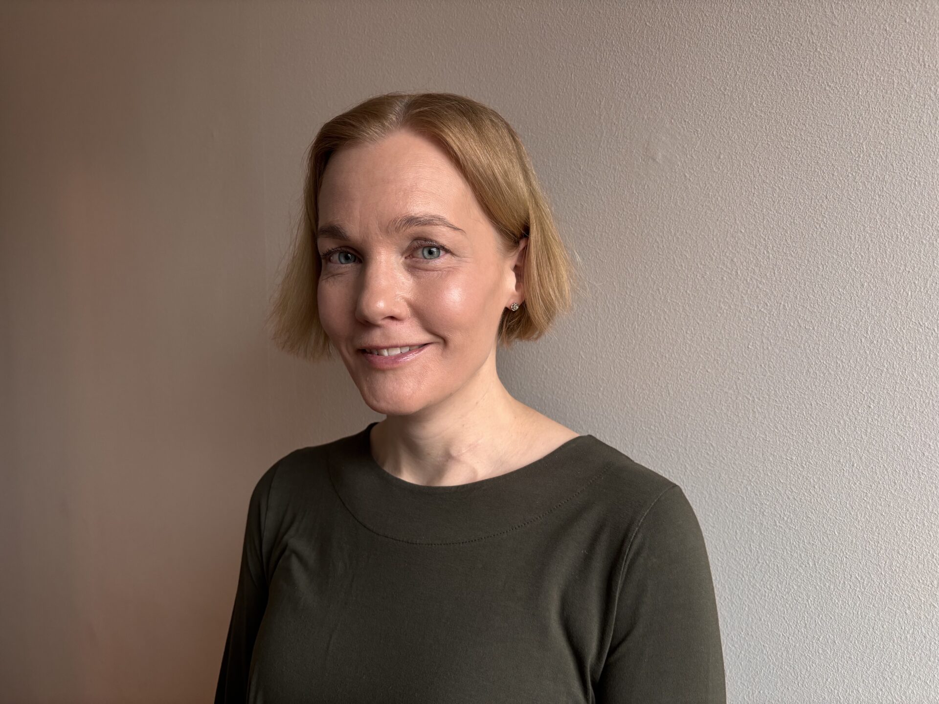 Nainen, jolla on vihreä paita ja vaalea lyhyt tukka, hymyilee kameralla vaaleaa taustaa vasten.