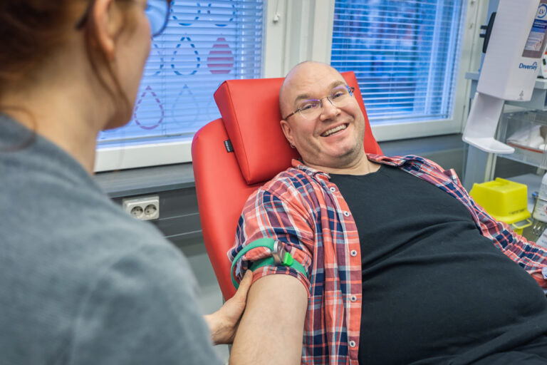 Mies hymyilee punaisella verenluovutustuolilla. Veripalvelun sairaanhoitajan profiili etualalla.