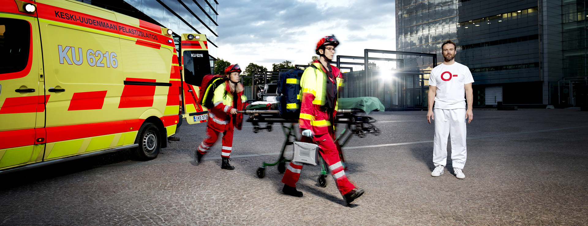 Sjuksköterskor rusar från ambulansen. En man i en vit t-shirt med O- i rött teckensnitt står bredvid honom.