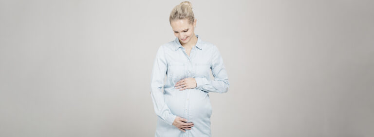 A pregnant woman wearing a light denim shirt gently holds her stomach.