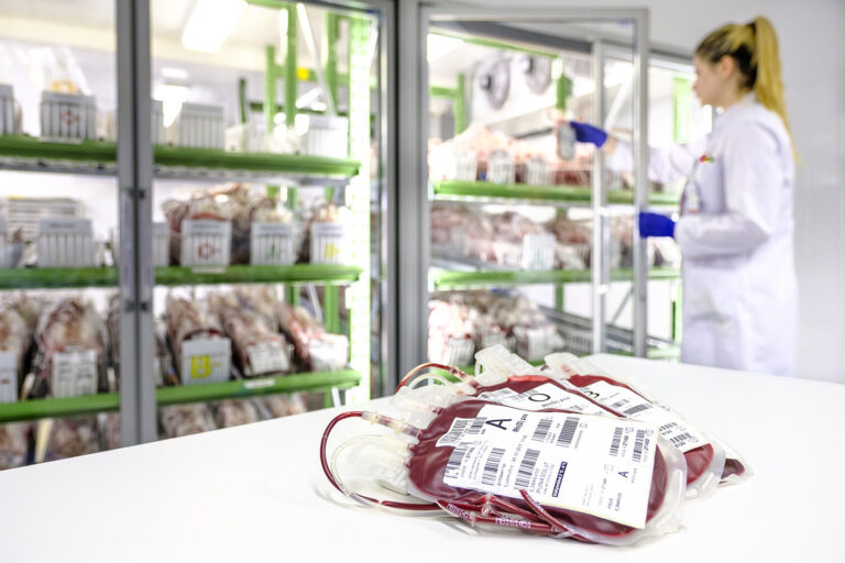 Red blood cell products on a table with a blood product warehouse and a Blood Service employee in a white coat in the background.