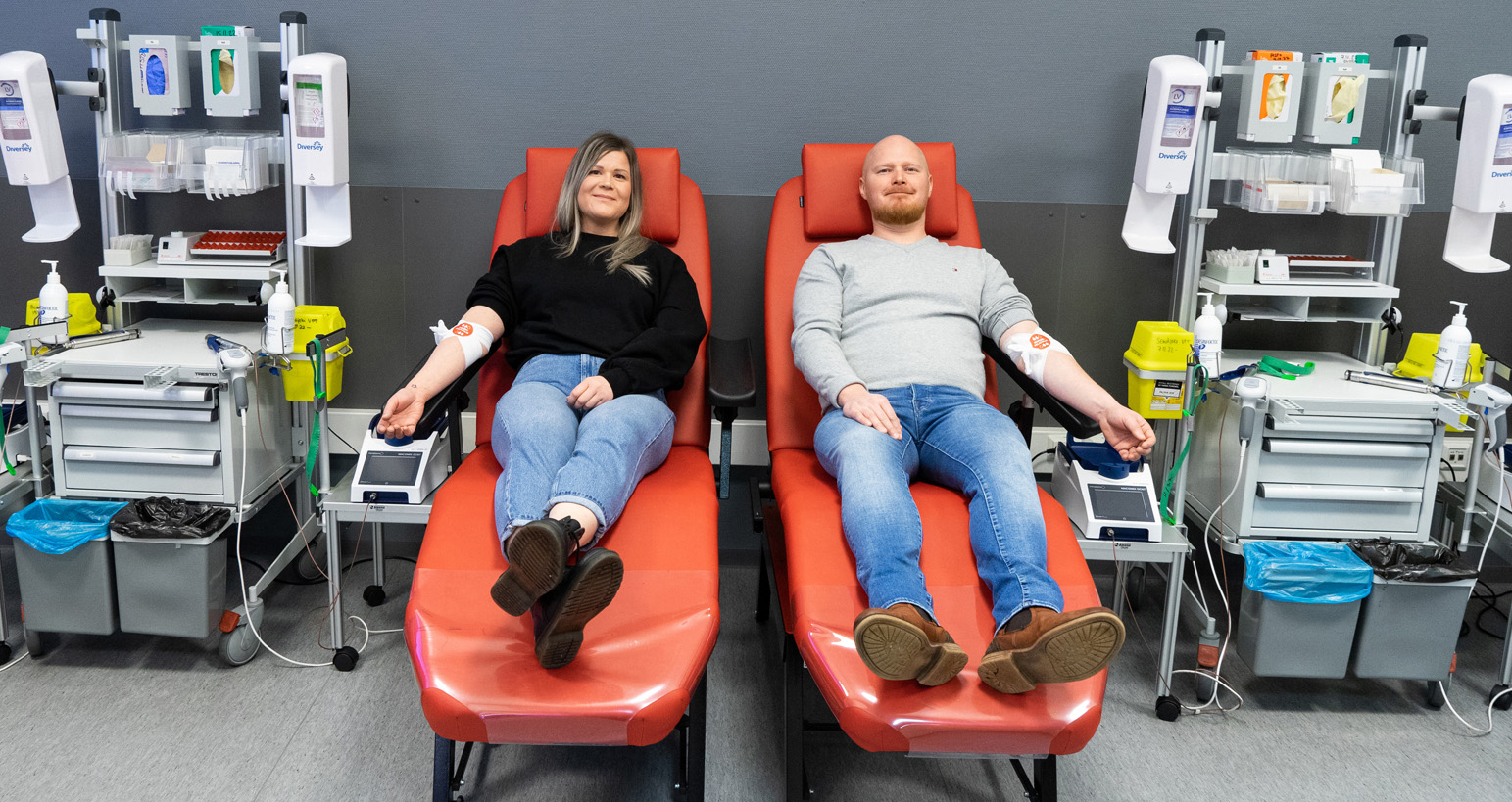 Kaksi verenluovuttajaa istuu vierekkäisillä verenluovutuspedeillä ja hymyilee kameralle.
