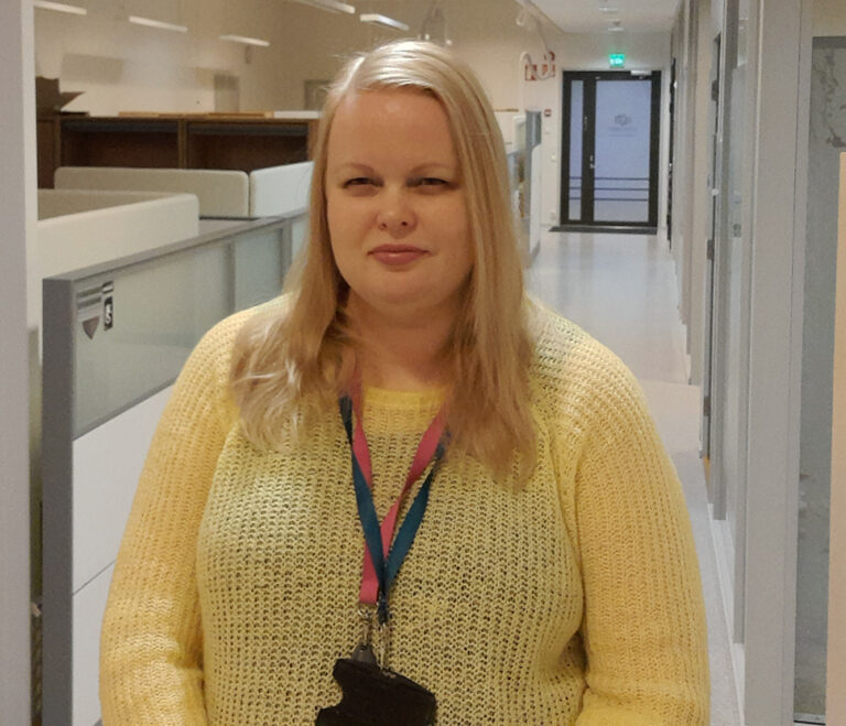 Nainen keltaisessa neuleessa, seisoo toimiston käytävällä.