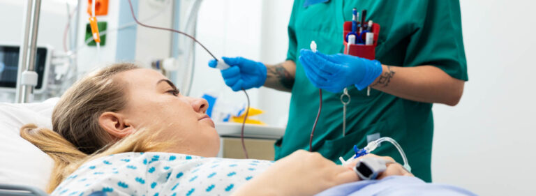 Nainen makaa sairaalasängyssä ja taustalla hoitaja valmistelee verensiirtoa.