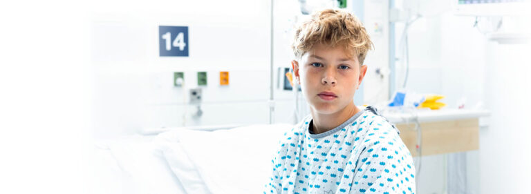 A young patient sits on the hospital bed in hospital clothes and looks into the camera.