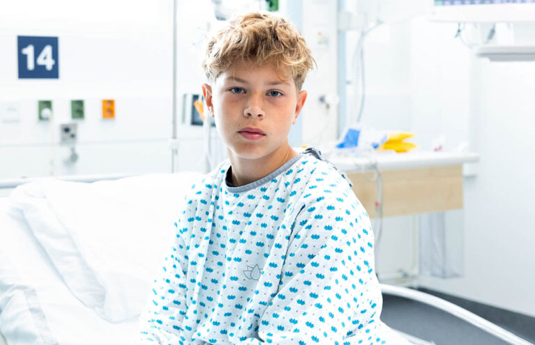 A young patient sits on the hospital bed in hospital clothes and looks into the camera.