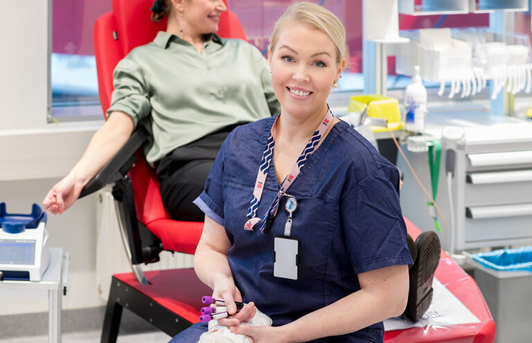Blodtjänsts sjuksköterska ler mot kameran, blodgivaren syns i bakgrunden.