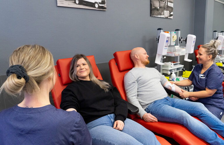 Two blood donors sit smiling while donating blood. Two nurses are sitting next to them.