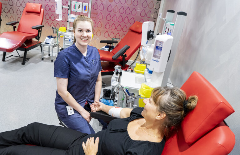 Blodtjänstsköterskan sitter bredvid blodgivaren vid donationsstället och tittar på kameran med ett leende.