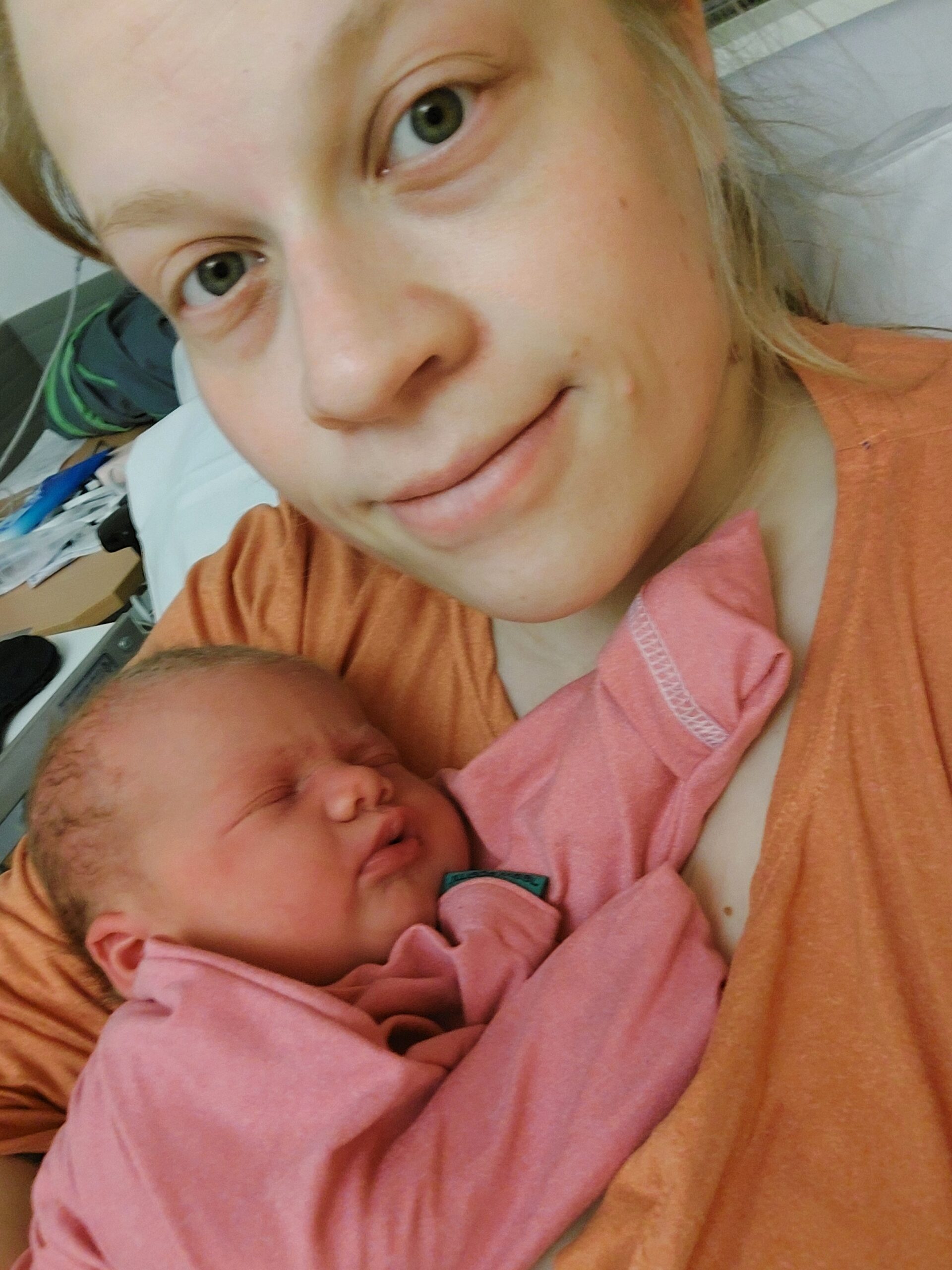 Henkilö hymyilee kameralle ja pitelee sylissään vastasyntynyttä vauvaa.