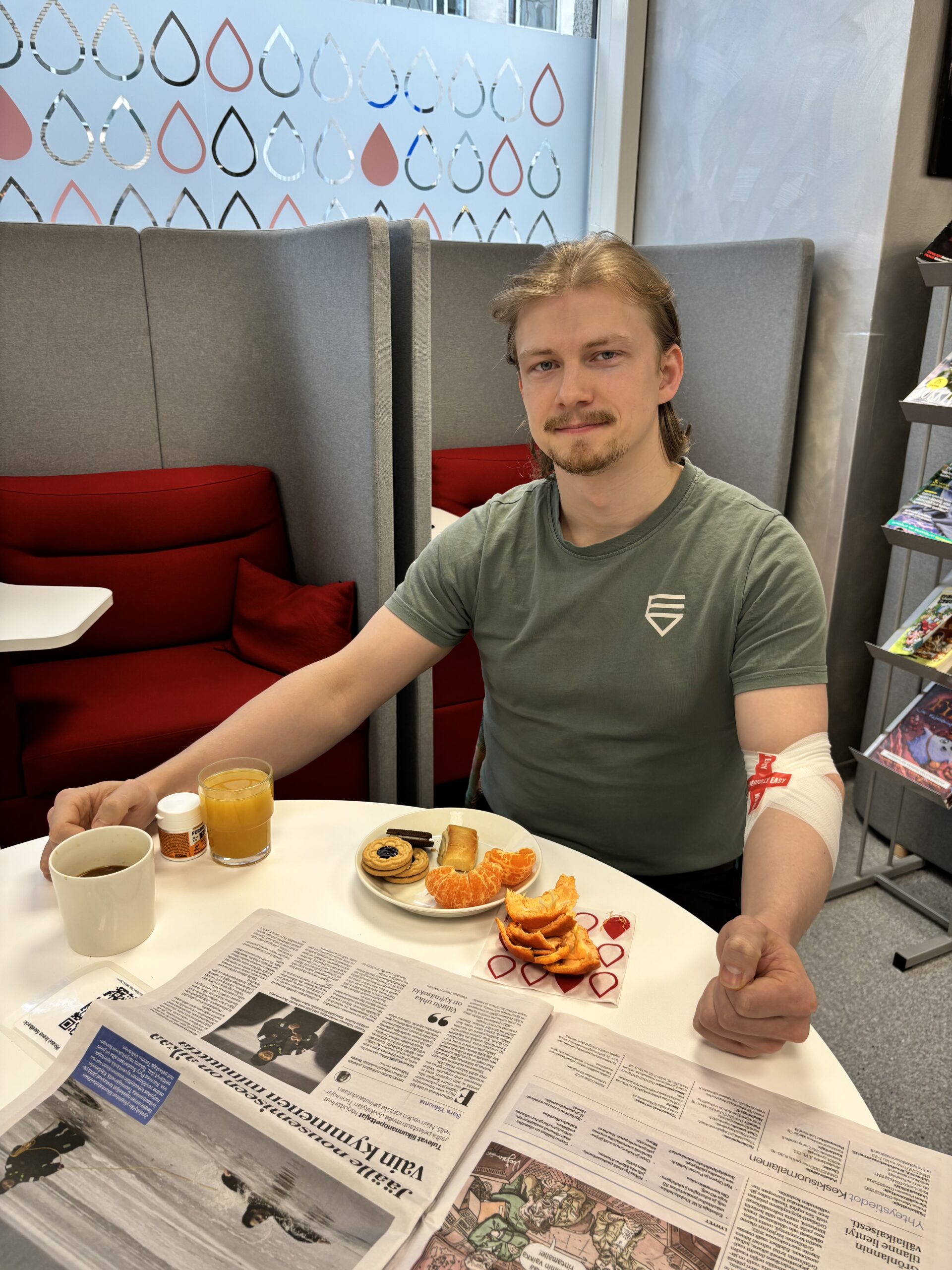 Mies jolla on vihreä t-paita istuu pöydän äärellä ja katsoo kameraan. Edessä on välipalaa ja sanomalehti.